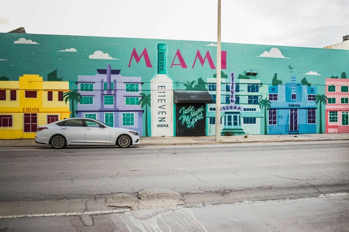 A colorful mural with large pink MIAMI letters and pastel Art Deco-style buildings painted on a wall, with a white car parked in front